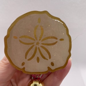 May include: A gold and white sand dollar ornament with a glitter finish. The sand dollar has a five-pointed star design in the centre.
