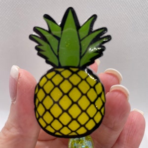 Könnte beinhalten: Ein gelber und grüner Ananas-Anhänger mit schwarzem Umriss. Die Ananas ist aus einem glänzenden, strukturierten Material gefertigt.