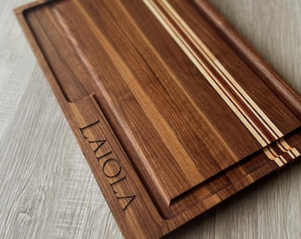 May include: A rectangular wooden cutting board with a dark brown finish. The board features a juice groove, a handle, and the engraved word "LAIOLA". Three parallel stripes of light wood and red accents adorn the top.