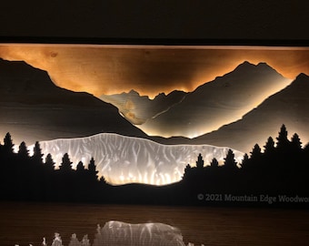 May include: A wooden shadow box with a mountain scene illuminated by warm white lights. The mountains are cut out of wood and layered to create a 3D effect. The box is framed in black wood and has a light wood background. The text "2021 Mountain Edge Woodworks" is visible in the bottom right corner.