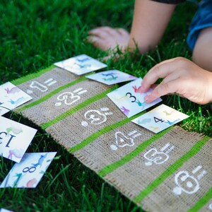 May include: A child plays a matching game on a burlap mat with green stripes. The game features colorful cards with numbers and animal illustrations. The child is placing a card with the number "3" on the mat.