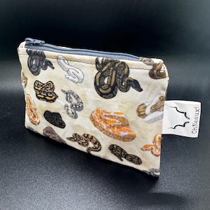 May include: A small, rectangular zippered pouch with a beige background and a repeating pattern of colorful snake illustrations. The pouch has a gray zipper and a white tag that reads "OhNeaux!"