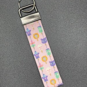 May include: A pink fabric keychain wristlet with a pattern of cartoon popsicles, skulls, and cat faces. The keychain has a silver metal clasp and key ring.