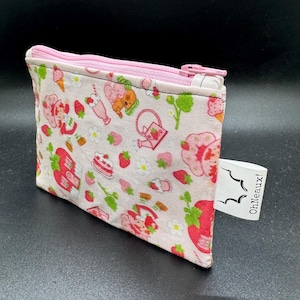 May include: A small, rectangular, zippered pouch with a pink zipper and a light pink background. The pouch is decorated with a pattern of strawberries, flowers, and a cartoon character. The tag reads "OhNeaux!"
