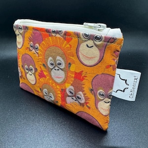 May include: A small, rectangular orange zippered pouch with a repeating pattern of cartoon orangutan faces in shades of brown, orange, and purple. The pouch has a white zipper and a white tag that reads "OhNeaux!"