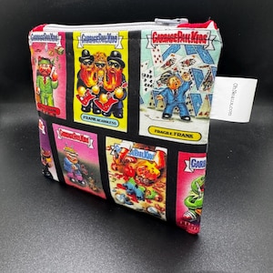 May include: A rectangular fabric zipper pouch featuring a colorful Garbage Pail Kids print with various character cards, including Frank Beardless and Fragile Frank. The pouch has a white side tag with the text OhNeaux.com printed in black ink.