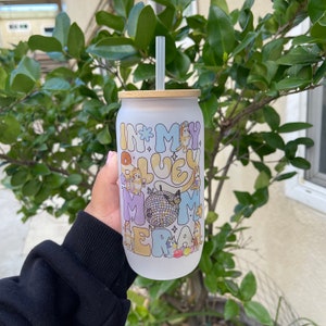 May include: Frosted glass tumbler with a bamboo lid and straw. The tumbler features the text "IN MY BLUEY MOM ERA" in pastel colors, along with cartoon characters, a disco ball, and a butterfly. The tumbler is held in front of a green leafy background.