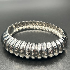 May include: A silver-colored bracelet with a ribbed design. The bracelet is made of metal and has a flexible, expandable design. The bracelet is circular and appears to be unworn.