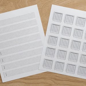 May include: Two white sheets of paper with musical notation. One sheet has horizontal lines for writing music, and the other has multiple guitar chord diagrams. The papers are on a light brown wooden surface.