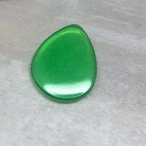 May include: A teardrop-shaped, translucent green object. The smooth surface reflects light, creating highlights. The object is set against a textured, light-colored background.