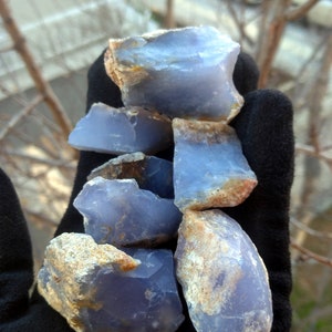 May include: A collection of rough, blue chalcedony stones with tan and brown edges. The stones are held in a black glove, with a blurred outdoor background.