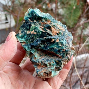 Può includere: Una pietra grezza di forma irregolare, con una miscela di tonalità verde acqua, verdi e marroni. La superficie ha un aspetto strutturato e cristallino, con aree di variazione cromatica intensa. La pietra è tenuta in mano, mostrando la sua forma naturale e grezza.