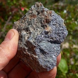 May include: A close-up of a dark gray rock with a metallic sheen. The rock has a rough, uneven surface and is covered in small, shiny crystals.