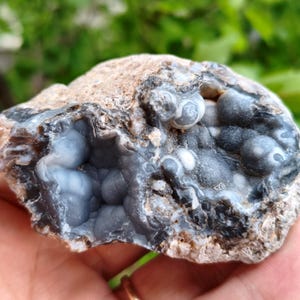 Puede incluir: Un espécimen mineral en bruto con una corteza exterior rugosa de color tostado que se abre hacia una cavidad llena de formaciones botrioidales lisas. El interior muestra tonos de gris translúcido, carbón oscuro y blanco con texturas redondeadas.