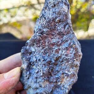 Può includere: Una roccia ruvida di forma irregolare con una lucentezza metallica. La superficie della roccia mostra un mix di tonalità marrone scuro, blu e argento, con strutture cristalline visibili. La consistenza generale appare ruvida e irregolare, suggerendo un campione minerale naturale.