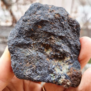 May include: A dark, textured rock with a rough surface. The rock has a mix of dark gray, black, and brown tones, with hints of green and orange. The natural stone appears to be a mineral specimen.