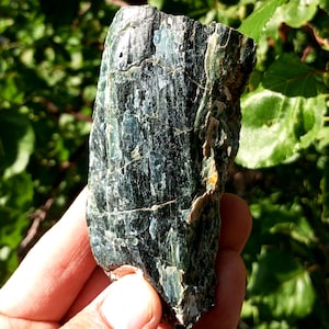 Könnte beinhalten: Ein grobes, dunkelgrünes Mineralexemplar mit einer geschichteten, strukturierten Oberfläche. Der Stein hat Streifen von hellgrün und blauen Farbtönen, mit natürlichen Rissen und Unvollkommenheiten. Das Exemplar wird in einer Hand vor einem verschwommenen grünen Hintergrund gehalten.