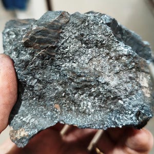 May include: A dark gray rock with a rough, textured surface. The rock has a layered appearance with visible crystalline structures and a metallic sheen. The overall color is a mix of dark gray and silver, with some areas appearing almost black.