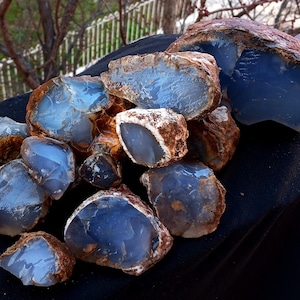 K&ouml;nnte beinhalten: Eine Sammlung roher, ungeschnittener blauer Chalcedonsteine. Die Steine haben ein durchscheinendes, glasartiges Aussehen mit einem hellblauen Farbton und braunen R&auml;ndern. Die Edelsteine werden auf einer schwarzen Oberfl&auml;che pr&auml;sentiert.