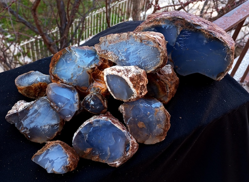 K&ouml;nnte beinhalten: Eine Sammlung roher, ungeschnittener blauer Chalcedonsteine. Die Steine haben ein durchscheinendes, glasartiges Aussehen mit einem hellblauen Farbton und braunen R&auml;ndern. Die Edelsteine werden auf einer schwarzen Oberfl&auml;che pr&auml;sentiert.