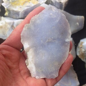 May include: A close-up of a light blue, translucent, irregularly shaped piece of chalcedony. The chalcedony is smooth and has a slightly cloudy appearance. It is being held in a hand.