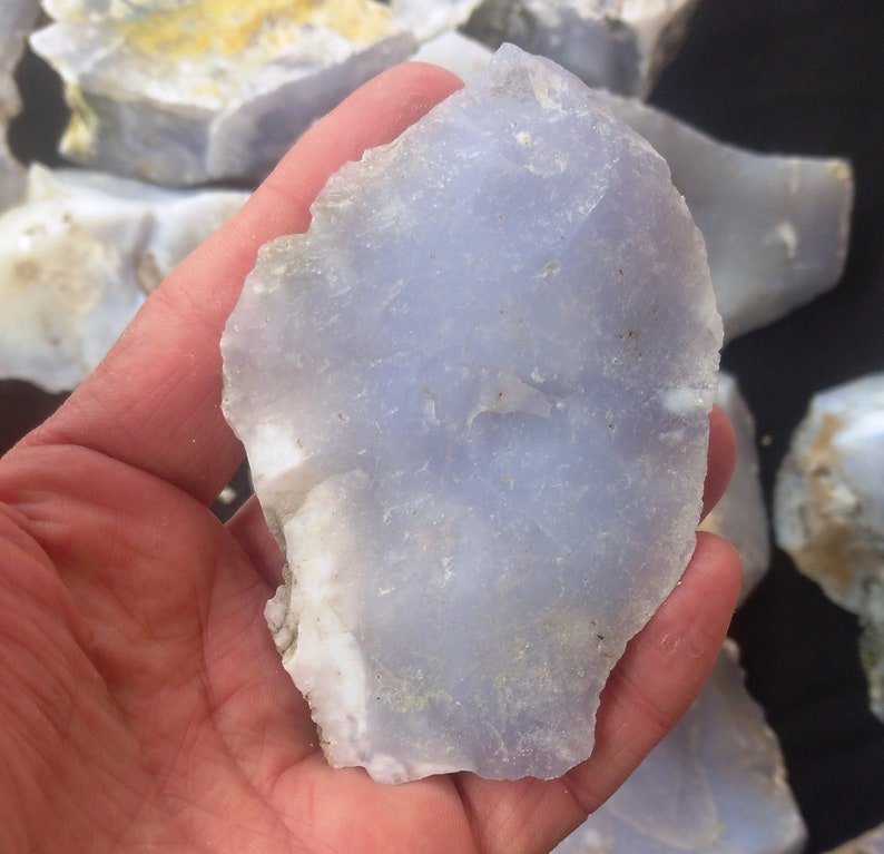 May include: A close-up of a light blue, translucent, irregularly shaped piece of chalcedony. The chalcedony is smooth and has a slightly cloudy appearance. It is being held in a hand.