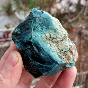 Può includere: Una mano tiene in mano un campione di minerale di colore turchese grezzo. La pietra presenta un mix di tonalità turchesi e marroni, con una superficie ruvida e bordi irregolari. Il colore del minerale è un turchese intenso e saturo, con aree di blu più scuro e marrone più chiaro.