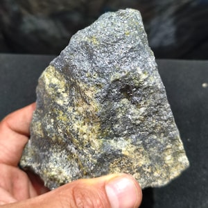 May include: A rough, irregularly shaped mineral specimen featuring a textured surface with a mix of dark gray, charcoal, and muted olive-green mineral deposits. The stone displays a crystalline, granular appearance with varied light-reflecting facets.