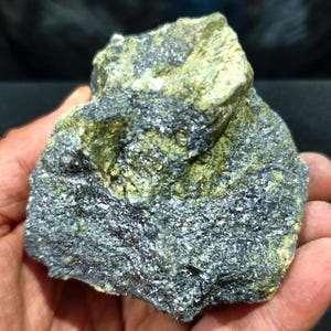 May include: A raw mineral specimen featuring a combination of metallic dark gray, crystalline textures and patches of matte, olive-green rock. The surface displays a rough, uneven topography with sparkling reflective facets throughout the dark sections.