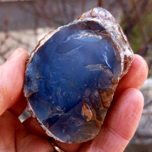 K&ouml;nnte beinhalten: Ein roher, nat&uuml;rlicher blauer Chalcedon-Edelstein. Der Stein hat eine tiefblaue Farbe mit Bereichen von hellem Blau und Braun. Die Kanten des Steins sind rau und uneben und zeigen die nat&uuml;rliche Bildung des Minerals. Der Edelstein wird in einer Hand gehalten.