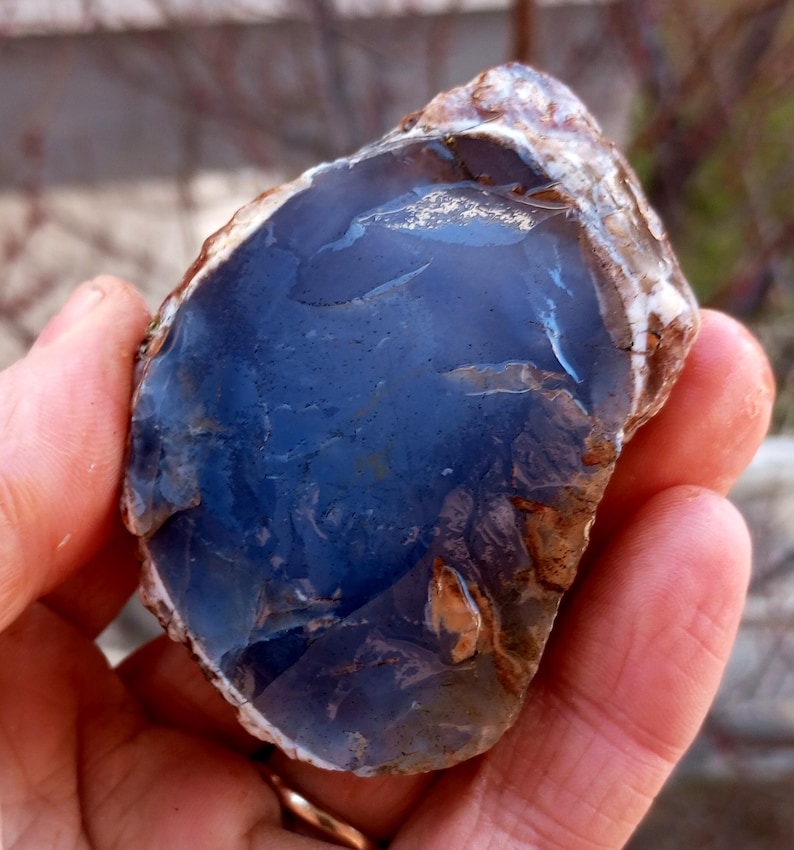 K&ouml;nnte beinhalten: Ein roher, nat&uuml;rlicher blauer Chalcedon-Edelstein. Der Stein hat eine tiefblaue Farbe mit Bereichen von hellem Blau und Braun. Die Kanten des Steins sind rau und uneben und zeigen die nat&uuml;rliche Bildung des Minerals. Der Edelstein wird in einer Hand gehalten.