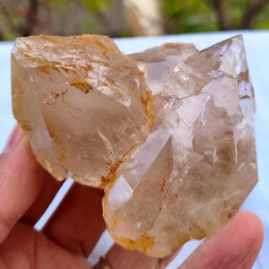 Pode incluir: Um aglomerado de cristais de quartzo transparente com uma tonalidade castanha clara e estruturas internas visíveis. Os cristais têm bordas afiadas e uma superfície áspera e texturizada. O quartzo é segurado em uma mão, mostrando sua forma natural.