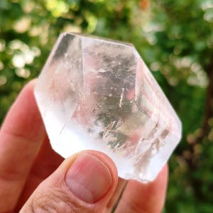 Könnte beinhalten: Ein klarer, facettierter Quarzkristall in sechseckiger Form mit glatten, polierten Oberflächen. Der Stein ist transparent und weist sichtbare interne Einschlüsse sowie natürliche Risse auf, die das Licht durch seine geometrische Struktur brechen.