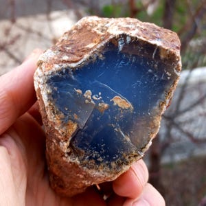 K&ouml;nnte beinhalten: Ein roh geschnittener Edelstein mit tiefblauem Innenraum und brauner, strukturierter Au&szlig;enseite. Der Stein wird in einer Hand gehalten, was seine nat&uuml;rliche, rohe Sch&ouml;nheit zeigt. Der blaue Innenraum hat ein glattes, poliertes Aussehen, das mit der rauen &auml;u&szlig;eren Schale kontrastiert.