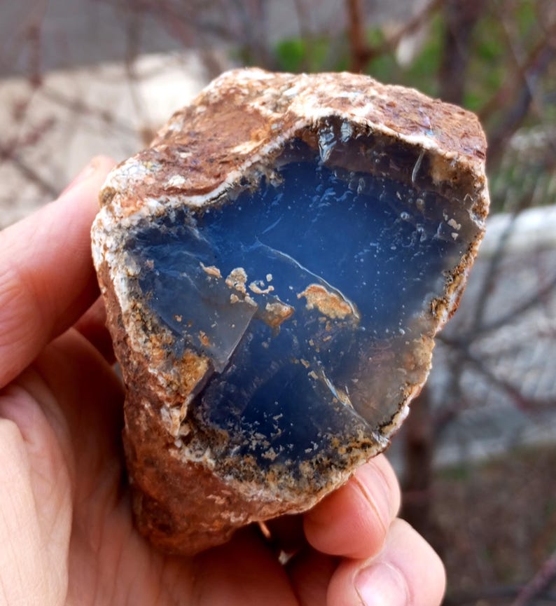 K&ouml;nnte beinhalten: Ein roh geschnittener Edelstein mit tiefblauem Innenraum und brauner, strukturierter Au&szlig;enseite. Der Stein wird in einer Hand gehalten, was seine nat&uuml;rliche, rohe Sch&ouml;nheit zeigt. Der blaue Innenraum hat ein glattes, poliertes Aussehen, das mit der rauen &auml;u&szlig;eren Schale kontrastiert.