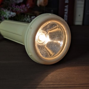 May include: A vintage, off-white flashlight with a bright, focused beam of light. The flashlight has a rounded head and a cylindrical body, with a small strap attached. The light is illuminating the surrounding area on a dark wooden surface.