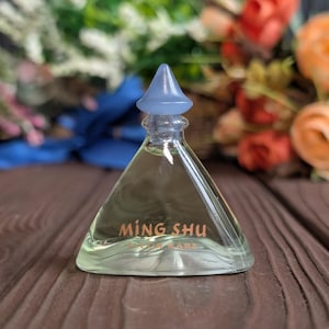 May include: A clear glass perfume bottle with a light blue stopper. The bottle has a triangular shape and contains a light yellow liquid. The words "MING SHU" are printed in gold on the front. The bottle is on a brown wooden surface.