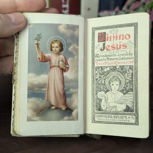 May include: Open miniature book with a color illustration of a child in a pink robe holding a dove and a sprig of leaves. The opposite page has text in Spanish, including "Divino Jesús" and "Editorial Regina Barcelona España."