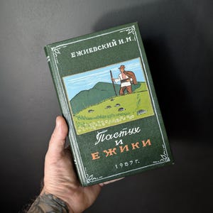 May include: A vintage hardcover book with a green cover, featuring a color illustration of a shepherd on a hillside. The title is in Russian, with the year 1987 printed below. The book is held in a hand against a dark background.