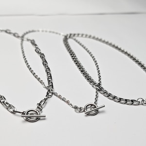 May include: Two silver chain necklaces with toggle clasps. The chains are different thicknesses and styles.