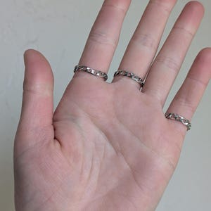 May include: A hand wearing three silver chain link rings. The rings are on the index, middle, and pinky fingers.
