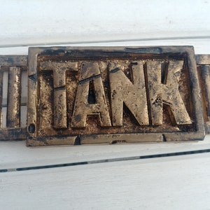 Puede incluir: Un letrero de metal dorado con la palabra "TANK" en relieve. El letrero tiene un aspecto desgastado y envejecido y está rodeado por un borde decorativo.