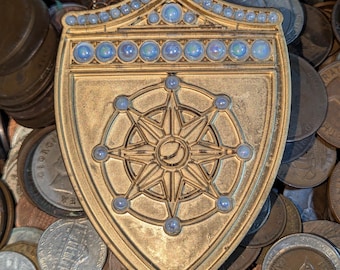 Puede incluir: Un colgante dorado en forma de escudo con un diseño de rosa de los vientos y detalles azules. El colgante, con una cadena, está rodeado de varias monedas, lo que sugiere un tesoro o un objeto de colección.