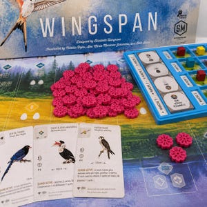 Könnte beinhalten: Ein Wingspan-Brettspielset mit einer großen Schachtel, einem Spielplan mit Wald- und Wiesenlandschaft, einem Haufen pinker blumenförmiger Futtermarker, drei Vogelkarten, einem blauen Kunststofftablett mit runden Markern sowie kleinen roten und gelben Würfeln.