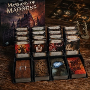 Könnte beinhalten: Ein Mansions of Madness Brettspiel-Set mit einer Schachtel, die ein dunkles Herrenhaus zeigt, mehreren Kartenstapeln in einem schwarzen Kunststoffhalter und zwei kleinen grauen Spielfiguren auf einer Holzoberfläche.