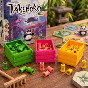 Könnte beinhalten: Die Takenoko-Spieleschachtel steht hinter drei rechteckigen Kunststoffkisten in Grün, Pink und Gelb. Jede Kiste enthält kleine zylindrische Bambus-Spielsteine in der jeweiligen Farbe. Lose Bambus-Spielsteine und ein Holzrechen liegen bereit.