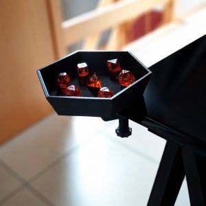 May include: A black hexagonal dice tray with a clamp mechanism attached to the edge of a table. The tray contains seven translucent red polyhedral dice with white numbers, including a d4, d6, d8, d10, d12, d20, and a percentile die.