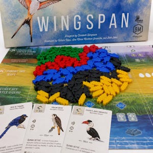 Op de afbeelding: De Wingspan bordspeldoos staat achter een speelbord met stapels groene, rode, blauwe, zwarte en gele voedselfiches in de vorm van vissen, zaden en knaagdieren. Drie vogelkaarten met illustraties worden op de voorgrond weergegeven.