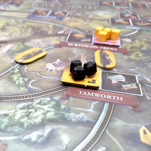 May include: A close-up of a board game with a yellow and brown game board. The board has a black train track and a yellow and brown train station. There are three black coal pieces on the board.