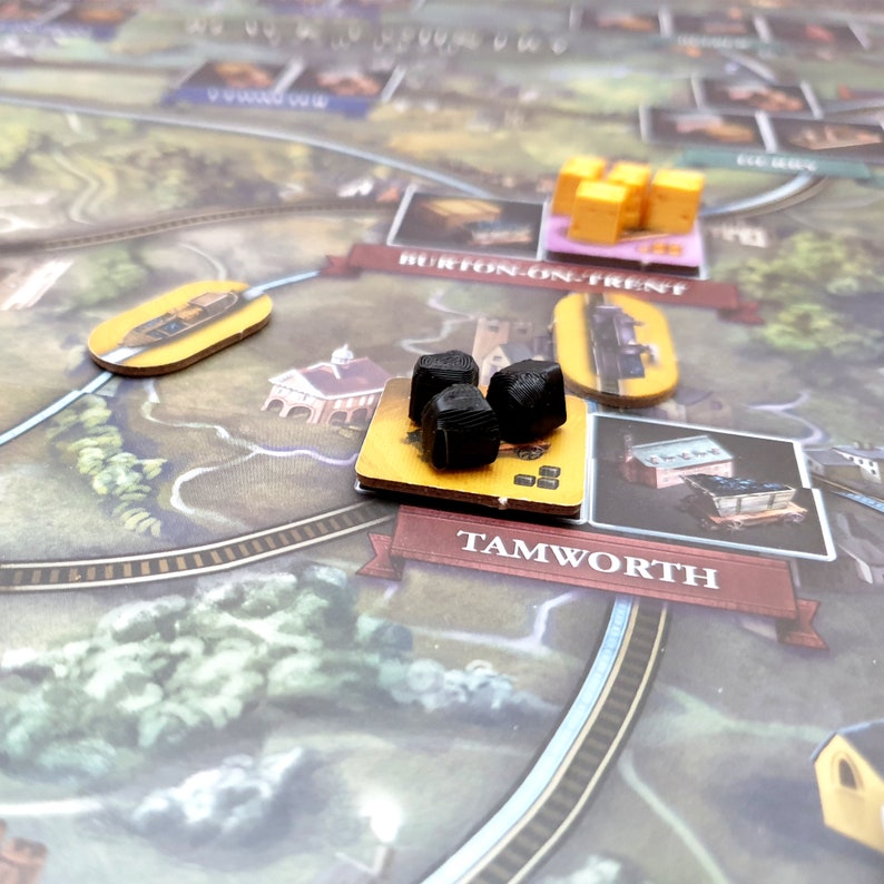 May include: A close-up of a board game with a yellow and brown game board. The board has a black train track and a yellow and brown train station. There are three black coal pieces on the board.